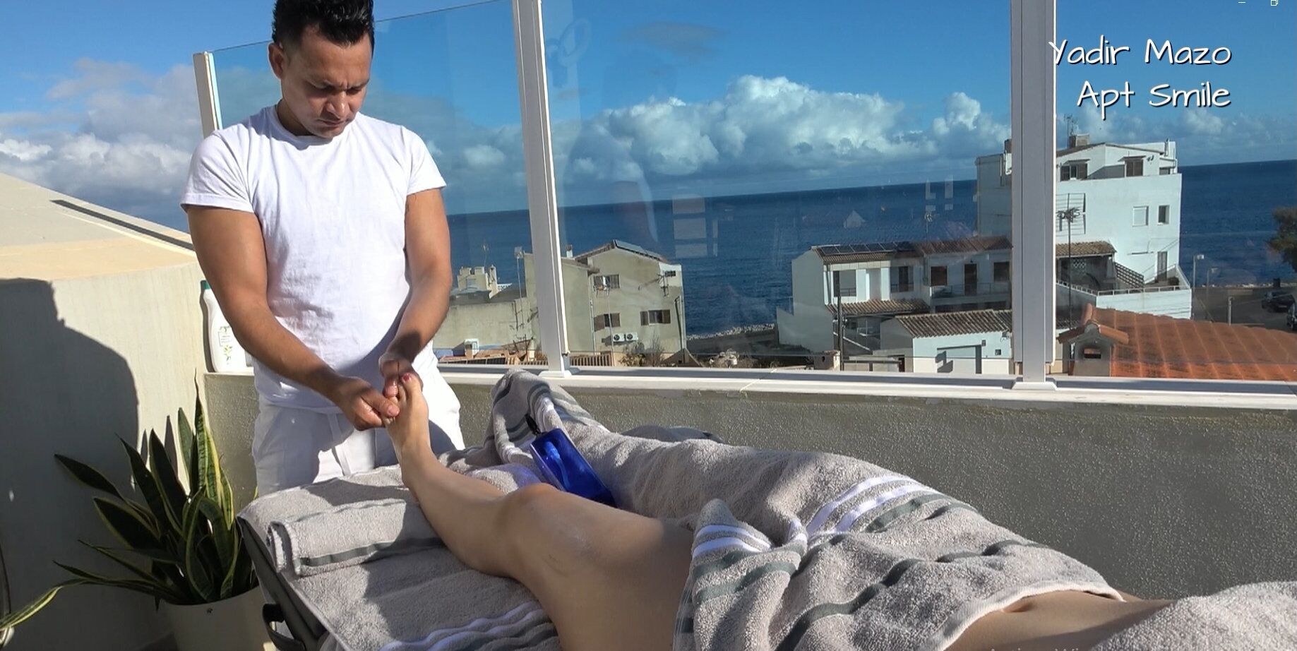Une personne reçoit un massage des pieds sur un balcon avec vue sur la mer et les bâtiments.