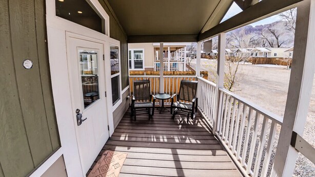 A covered porch with a wooden floor, a white door, and a view of a residential area. - Moab Tiny Home Near Arches | Sleeps 6 | Private Porch | Sand Flats (Moab)