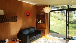 A modern living room with a black leather sofa, wooden shelves, and a large window offering a view of a garden. - Harbour Escape - tiny home in Lyttelton with Port Views (Lyttelton)