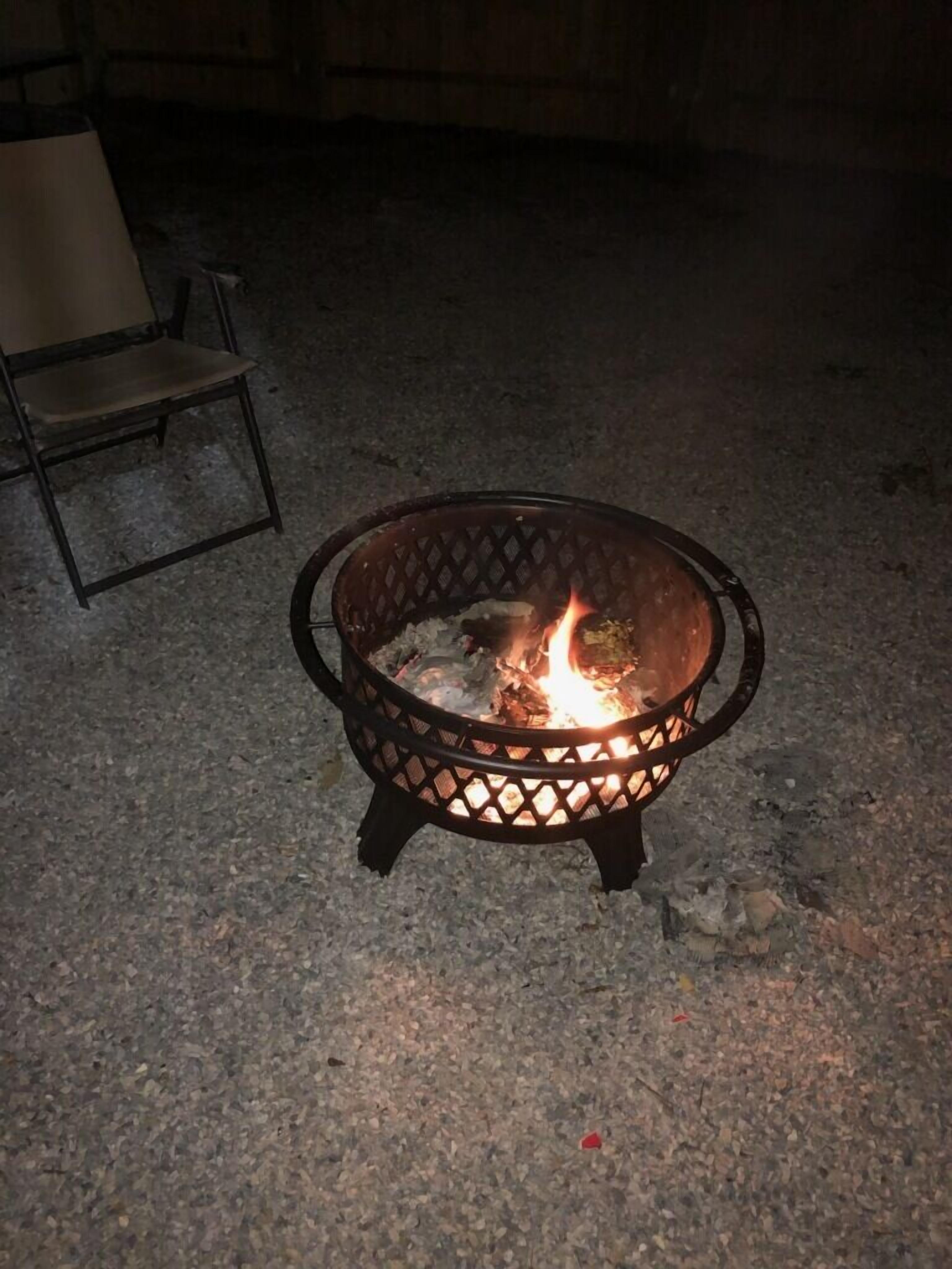 A metal fire pit with a fire burning inside, placed on a gravel surface.