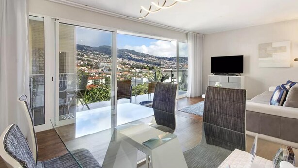 Una sala de estar moderna con una mesa de comedor de vidrio, asientos cómodos, un televisor de pantalla plana y un balcón con vistas a la ciudad.