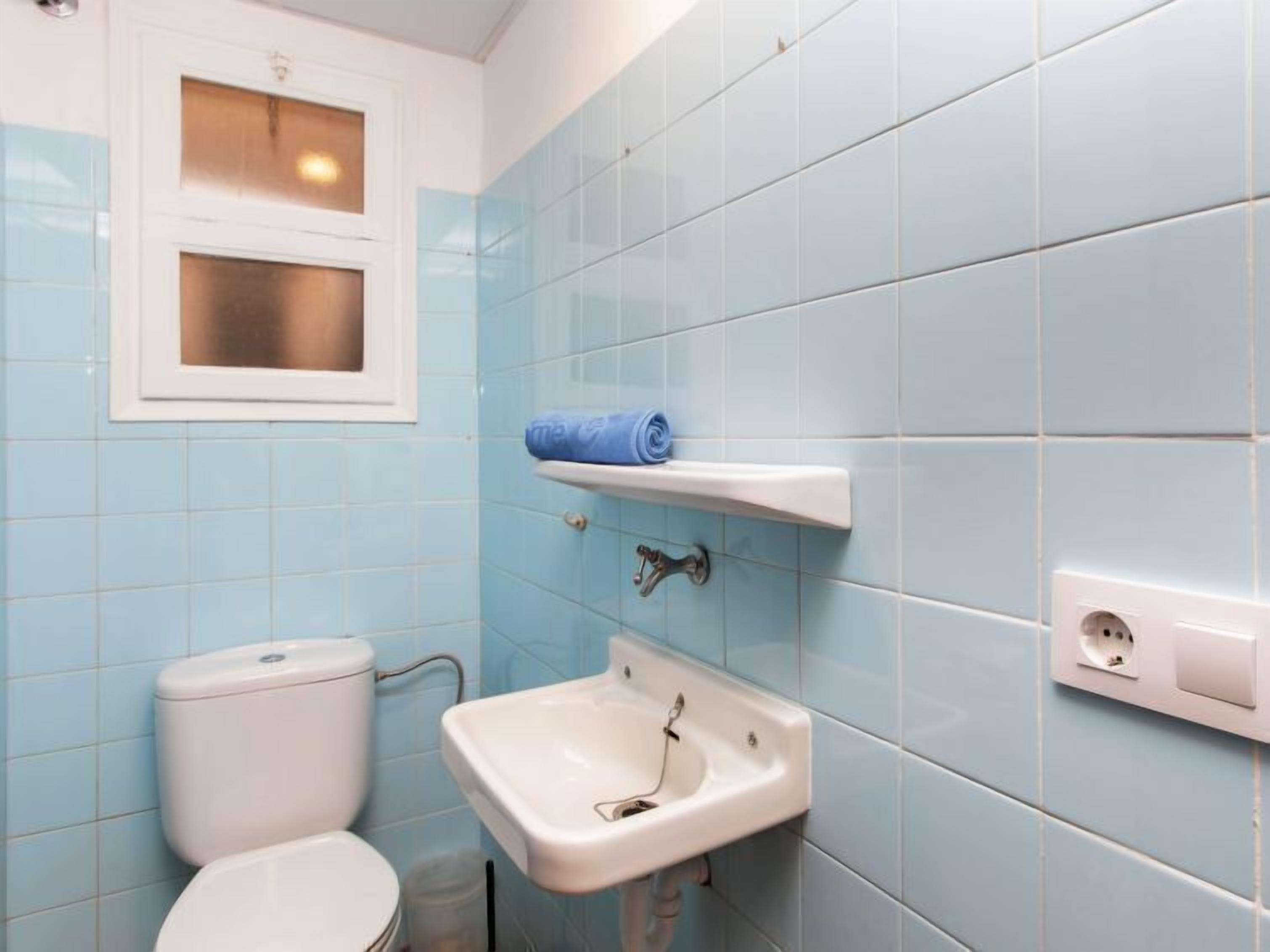 Une salle de bain avec des carreaux bleu clair, des toilettes, un lavabo et une fenêtre située au-dessus des toilettes.