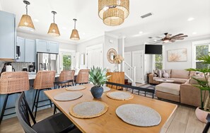 A modern kitchen with light blue cabinets, a dining area with a wooden table and woven placemats, and a living room with a beige sofa and a ceiling fan.