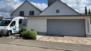 Une maison à deux étages avec un garage, un camping-car garé et un petit jardin agrémenté de plantes en pot.