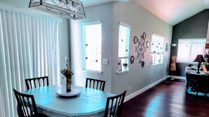 A dining area with a round table, chairs, and a vase of flowers. There are windows with blinds and wall decorations. - Huge Peaceful & Cheerful Home (Redding)