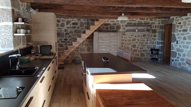 A modern kitchen with a central island, wooden cabinets, a stone wall, and a staircase.
