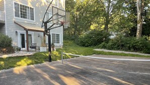 A basketball court with a net, a basketball hoop, and a house in the background.
