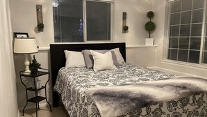 A bedroom with a bed, a nightstand, a lamp, and two windows with blinds.