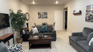 A living room with a sofa, armchair, coffee table, and a TV. There is a door leading to another room and a plant in the corner.