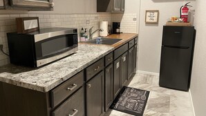 A kitchen with a microwave, sink, and refrigerator. There is a sign on the wall and a doormat on the floor.