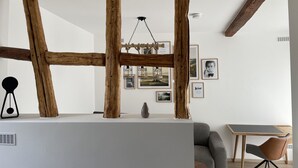Et værelse med synlige træbjælker, en hængende lampe, billeder i rammer på væggen, et bord og en sofa.