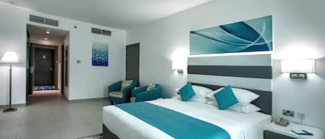 A modern hotel room with a large bed, a desk, and a chair. There is a painting on the wall and a lamp on the desk.