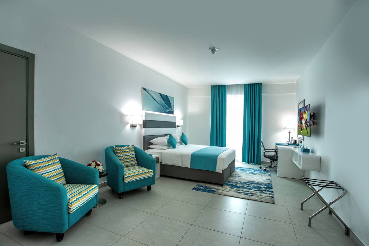 A modern hotel room with a bed, two armchairs, a desk, and a TV.