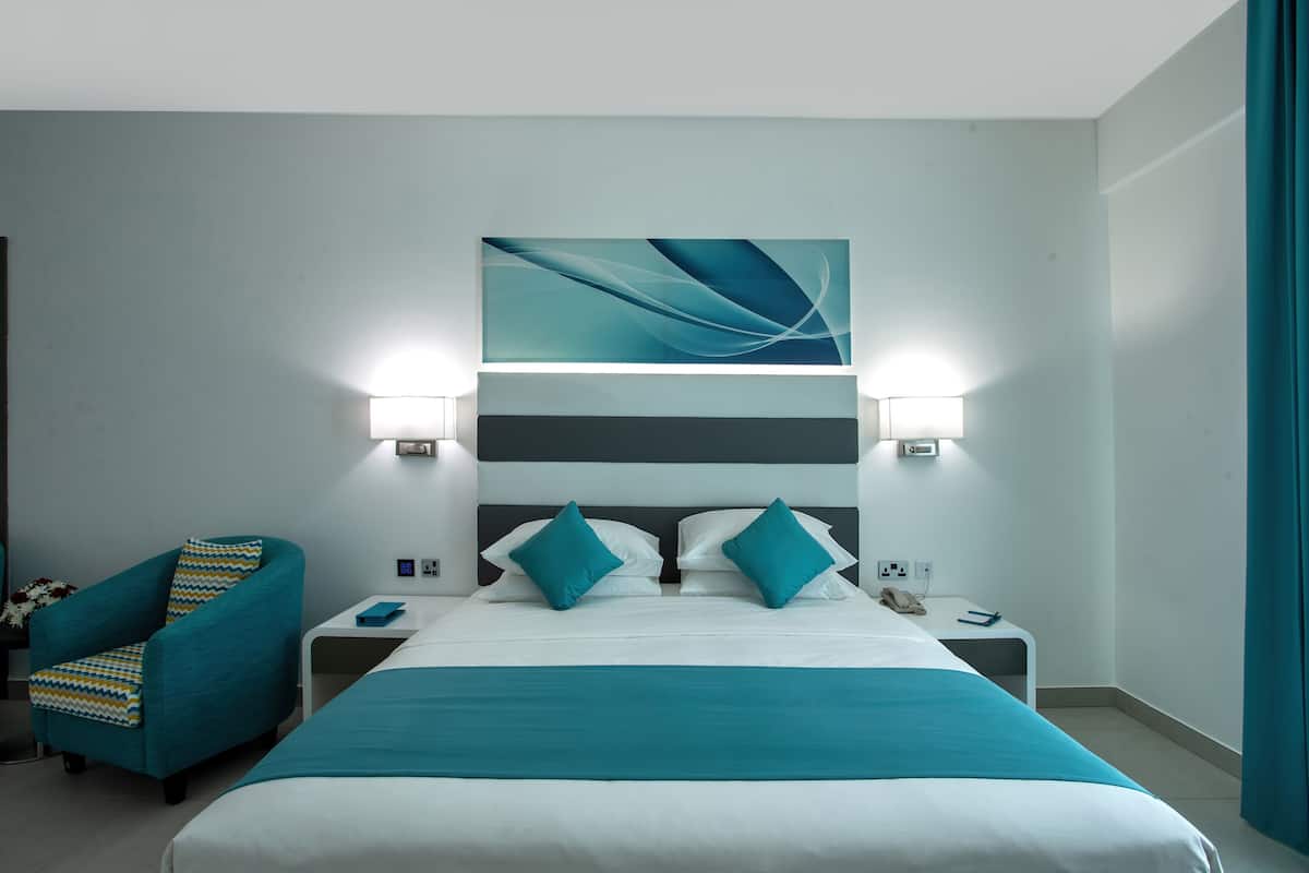 A modern hotel room with a large bed, two bedside tables, a chair, and a wall-mounted abstract painting.