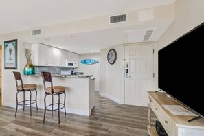 A kitchen with white cabinets, a microwave, and a clock on the wall. There are two bar stools and a large flat-screen TV on a wooden stand.