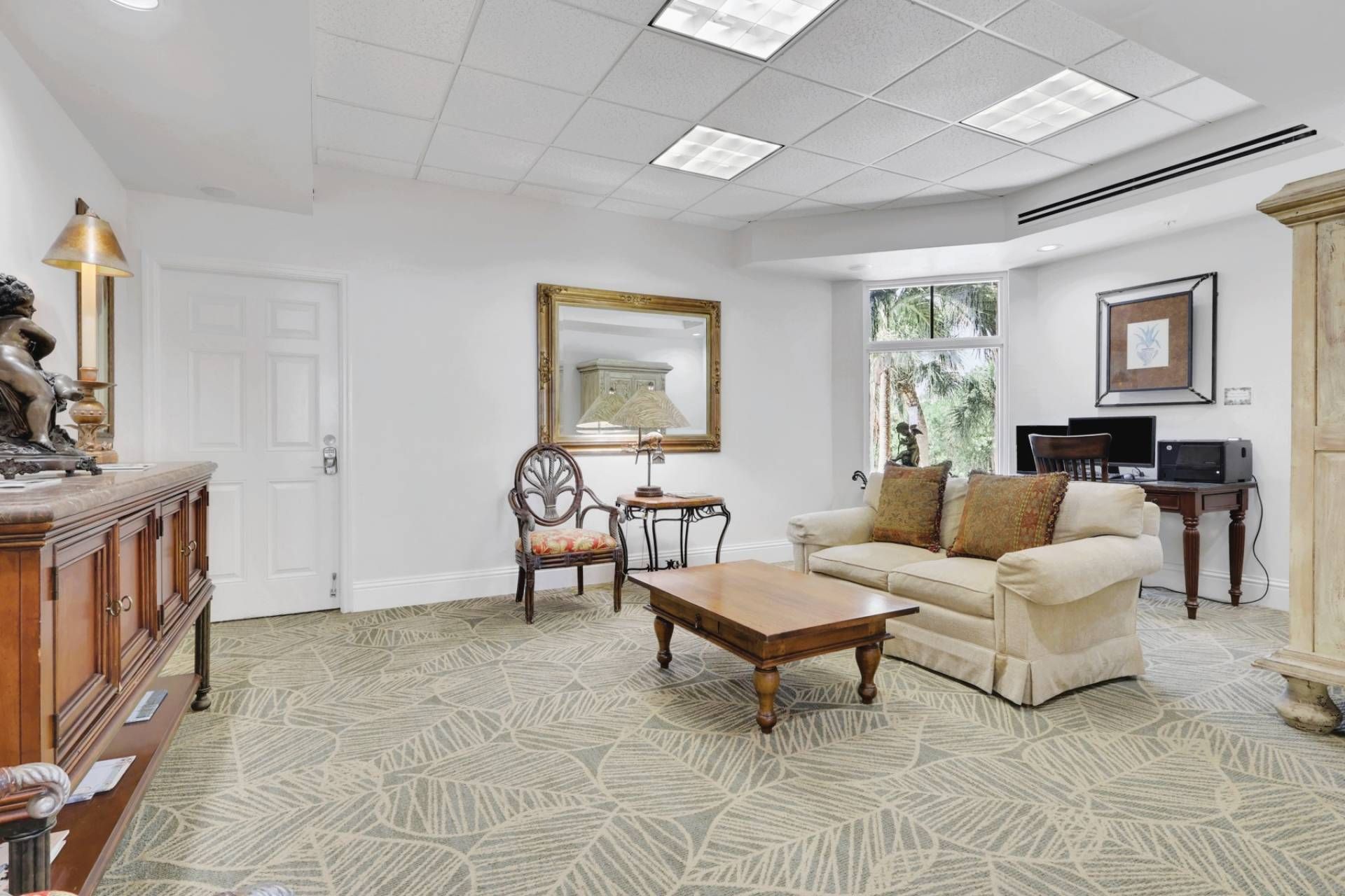 A living room with a sofa, coffee table, and a decorative chair. There is a fireplace, a television, and a window with a view of greenery.