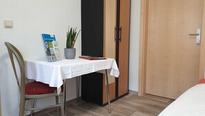 Habitació petita amb un llit, una taula amb estovell blanc, una cadira, una planta en test i una porta.