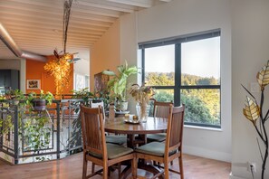 A dining area with a round table and chairs, surrounded by potted plants and a view of trees through a large window.