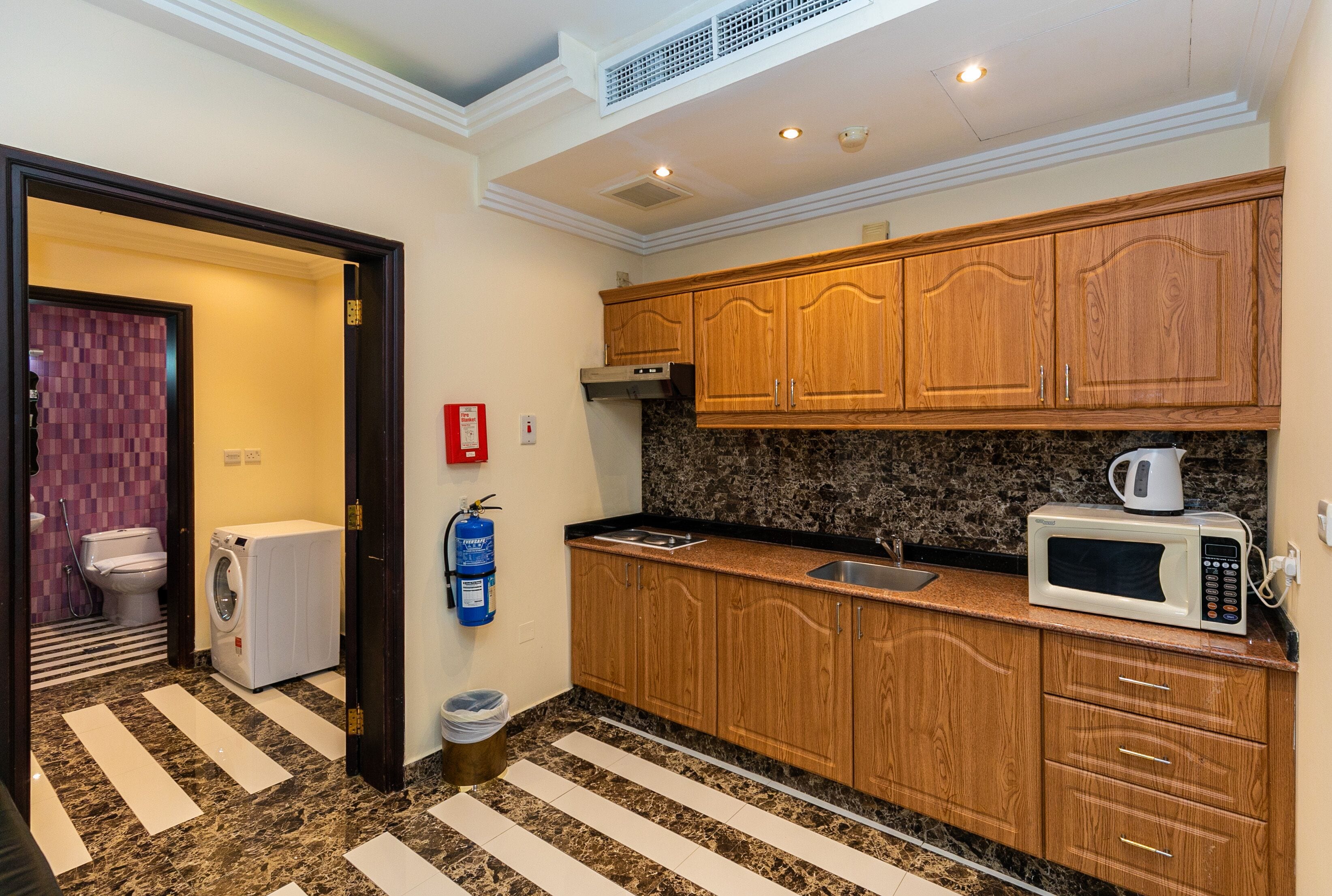 A compact kitchen with wooden cabinets, a microwave, and a built-in oven. A laundry machine is visible in the adjacent room.