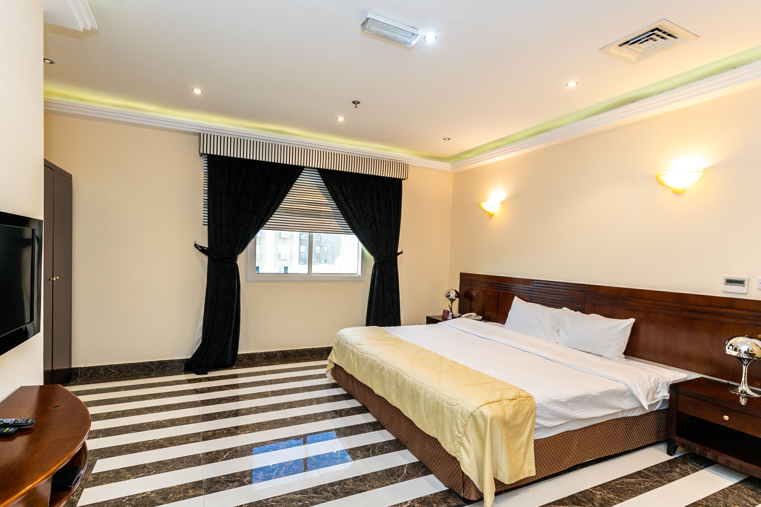 A hotel room with a large bed, a flat-screen TV, a wooden nightstand, and a window with curtains.