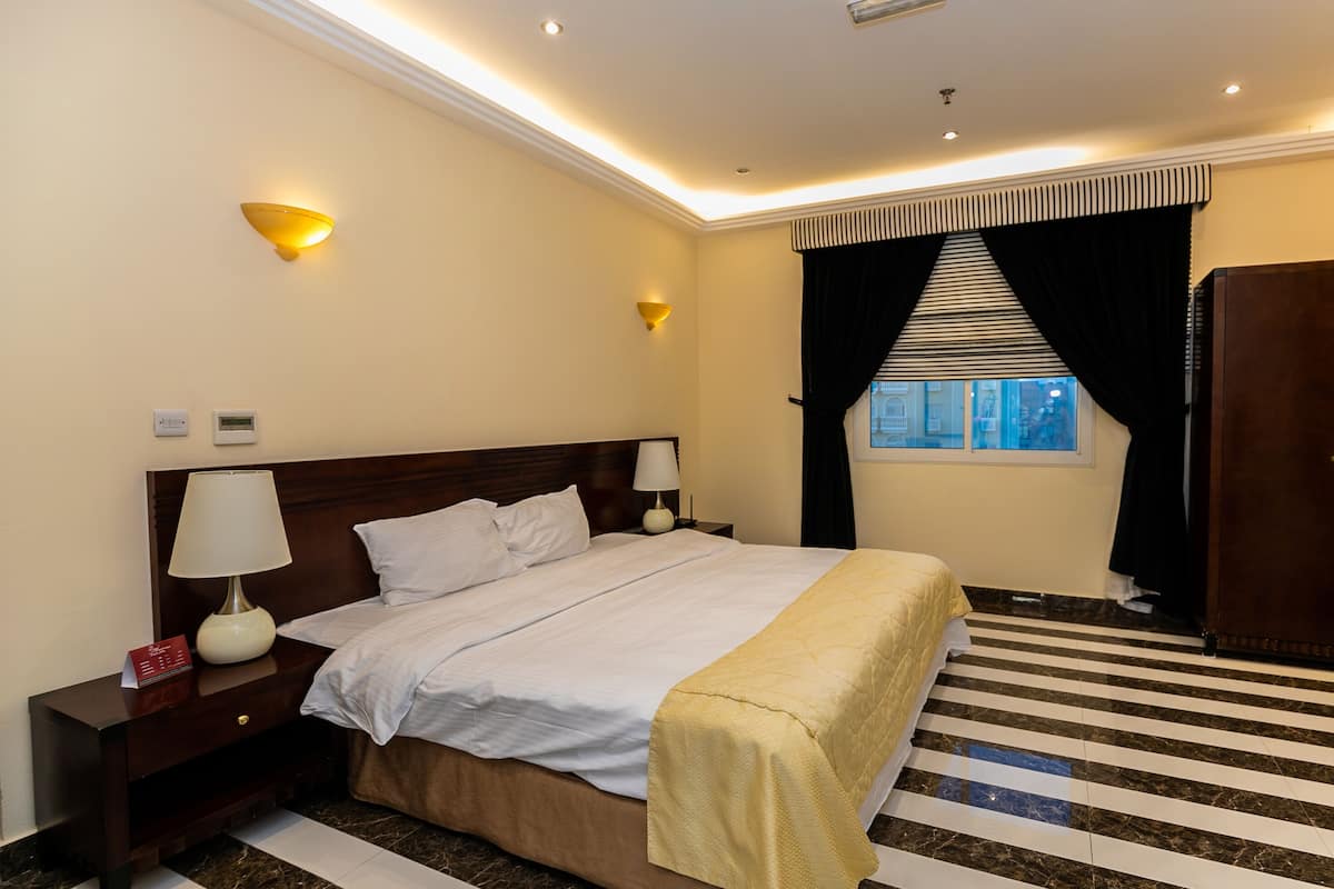 Une chambre d’hôtel avec un grand lit, deux tables de chevet avec des lampes, une fenêtre avec des rideaux noirs et une armoire en bois.