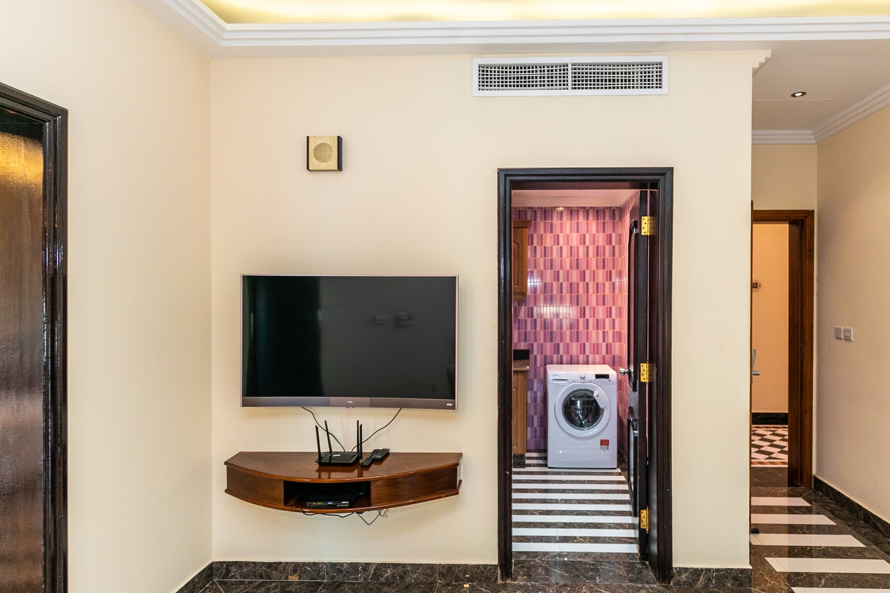 Une chambre d’hôtel moderne dotée d’un téléviseur à écran plat fixé au mur, d’un lave-linge et d’un sèche-linge intégrés, et d’un sol à motifs à carreaux.