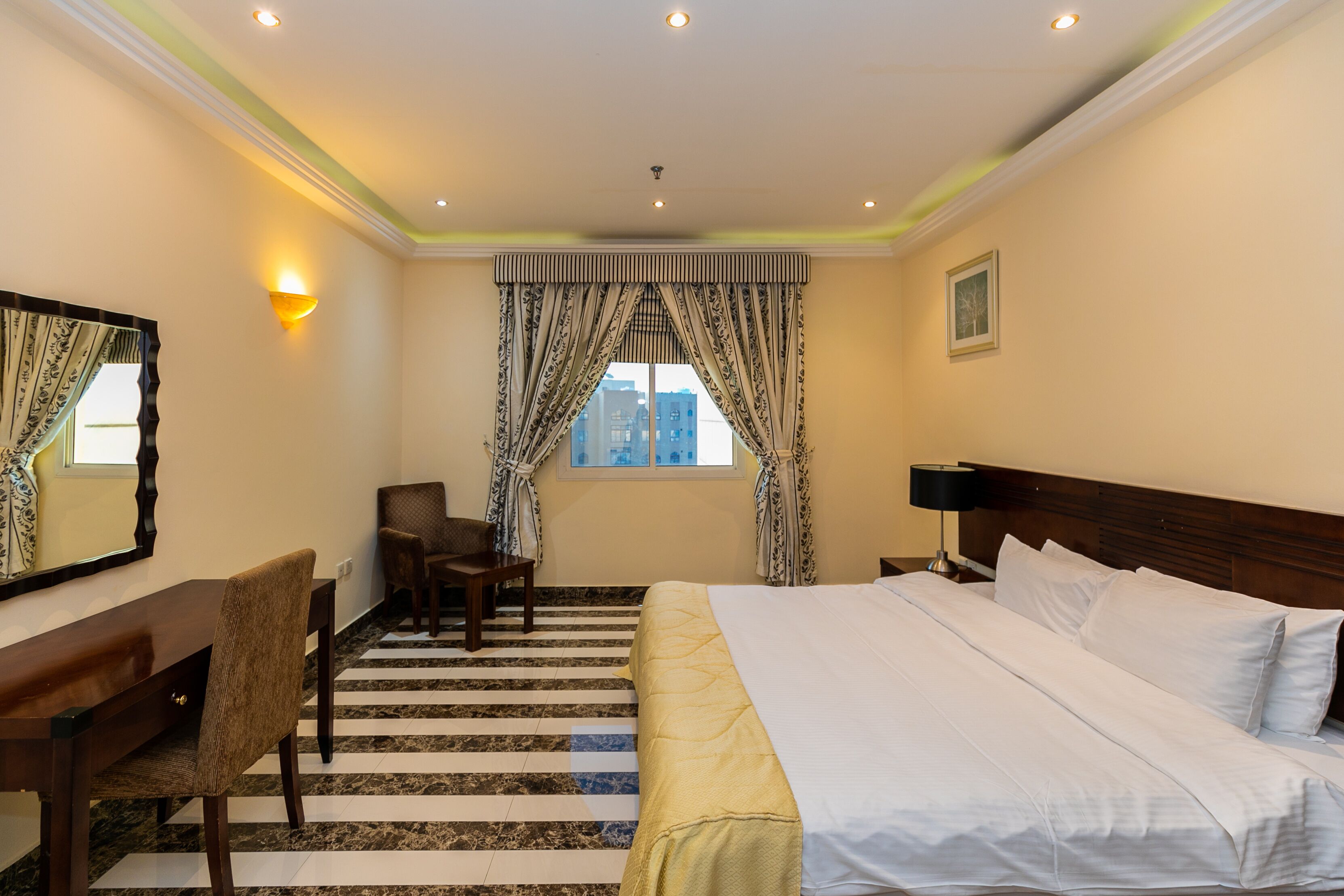 A hotel room with a bed, a desk, a chair, a window with curtains, and a nightstand with a lamp.