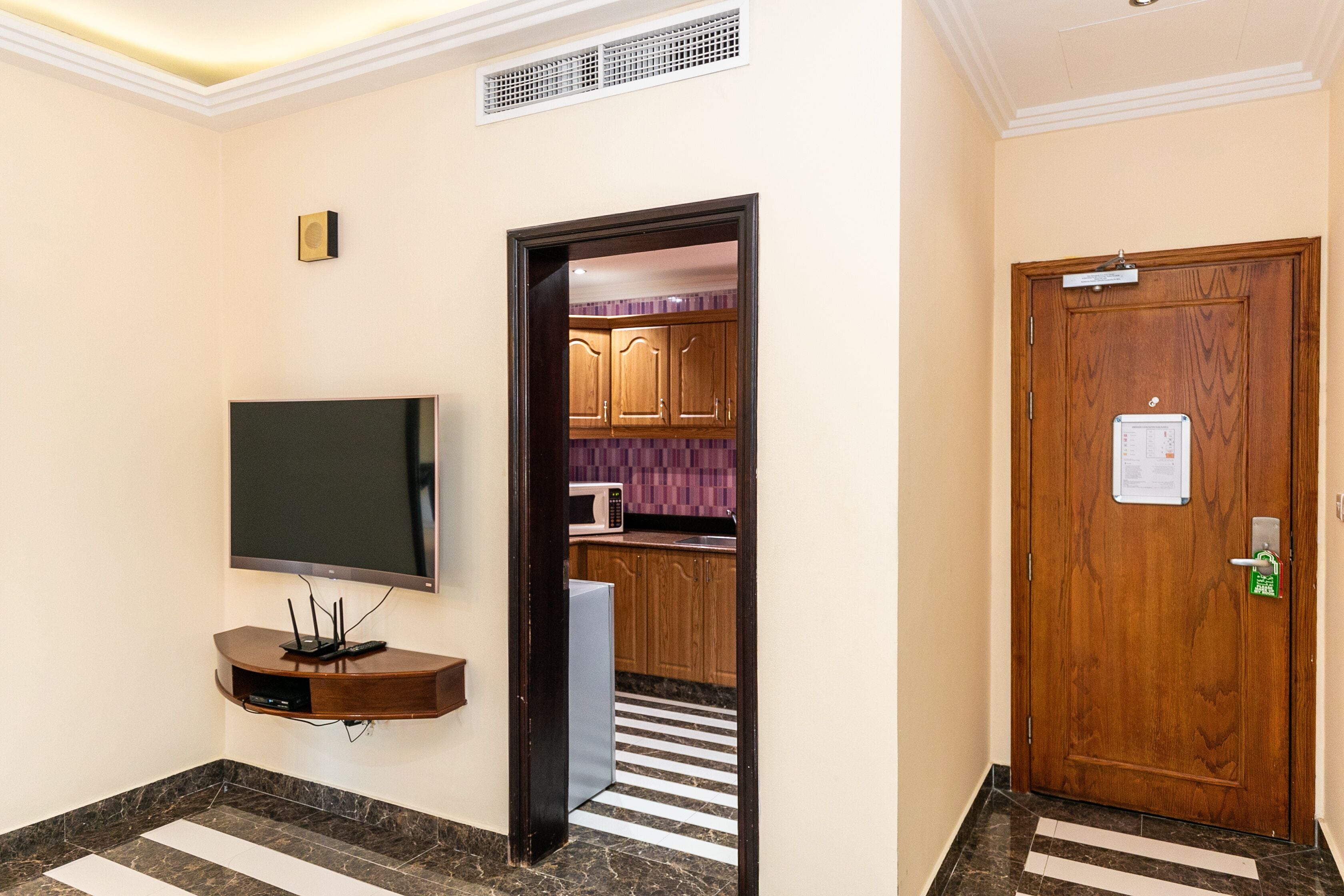A hotel lobby with a flat-screen TV mounted on a wooden console, a wooden door with a lock, and a striped floor leading to a kitchen area with wooden cabinets and a microwave.