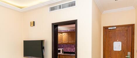 A hotel lobby with a flat-screen TV mounted on a wooden console, a wooden door with a lock, and a striped floor leading to a kitchen area with wooden cabinets and a microwave.