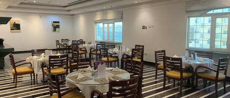 A spacious dining area with round tables and chairs, a striped floor, and large windows offering a view of the city.