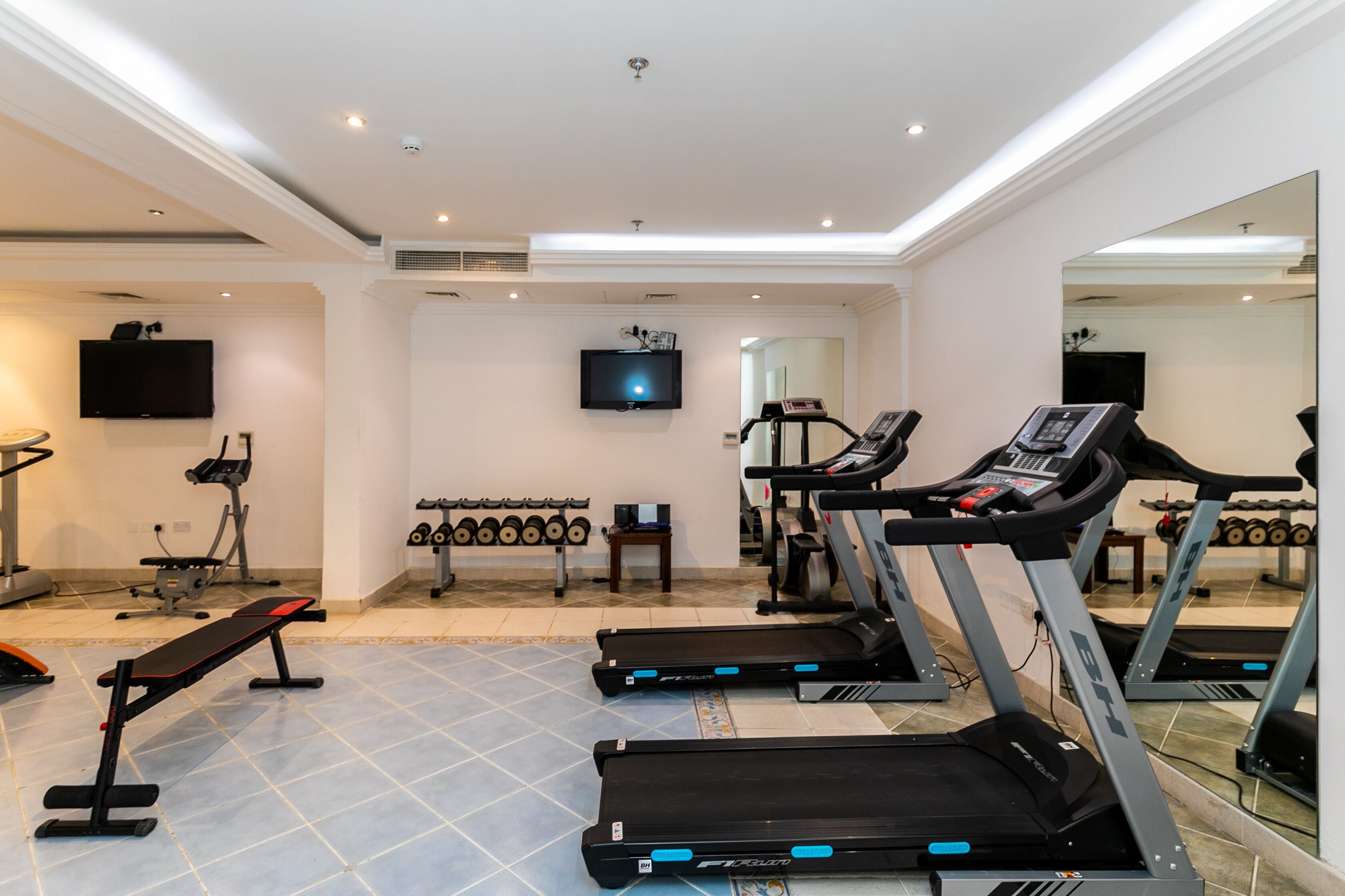 A modern gym with treadmills, a bench, dumbbells, and a TV.