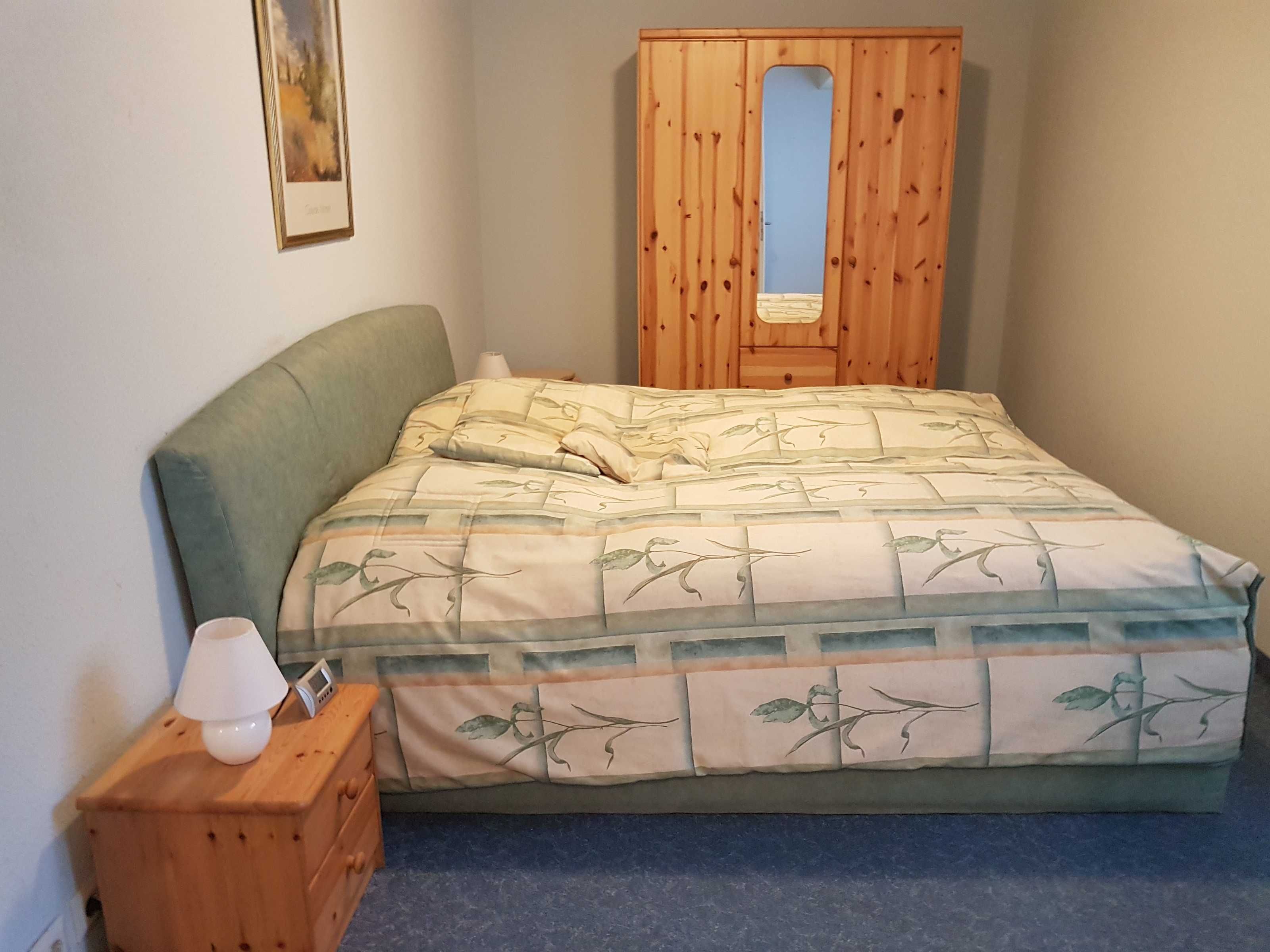 Een slaapkamer met een bed, een houten garderobe, een nachtkastje met een lamp en een ingelijst foto aan de muur.
