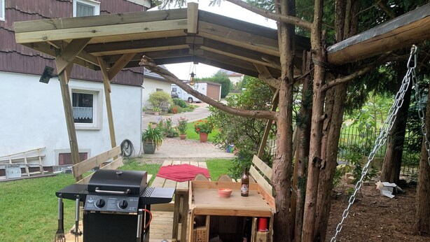 A wooden deck with a barbecue grill, a gas cylinder, and a wooden table with a red cloth. - Apartment Märzenbecher (Stolpen/OT Heeselicht)