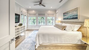 A bedroom with a large bed, a ceiling fan, a TV, and a window with a view of trees. - Amour La Mer by AvantStay | Rooftop Patio, Pool Access, Walk to Beach (Seacrest)
