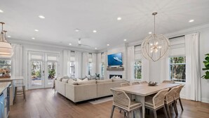 A spacious living room with a large beige sectional sofa, a dining area with a wooden table and wicker chairs, and a modern chandelier. - Amour La Mer by AvantStay | Rooftop Patio, Pool Access, Walk to Beach (Seacrest)