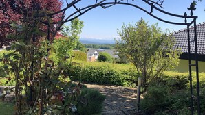 A stone pathway leading through a garden with a metal archway, surrounded by greenery and a view of distant houses. - Urlaub im Wunderschönen Hegau mit Alpenblick (Tengen)
