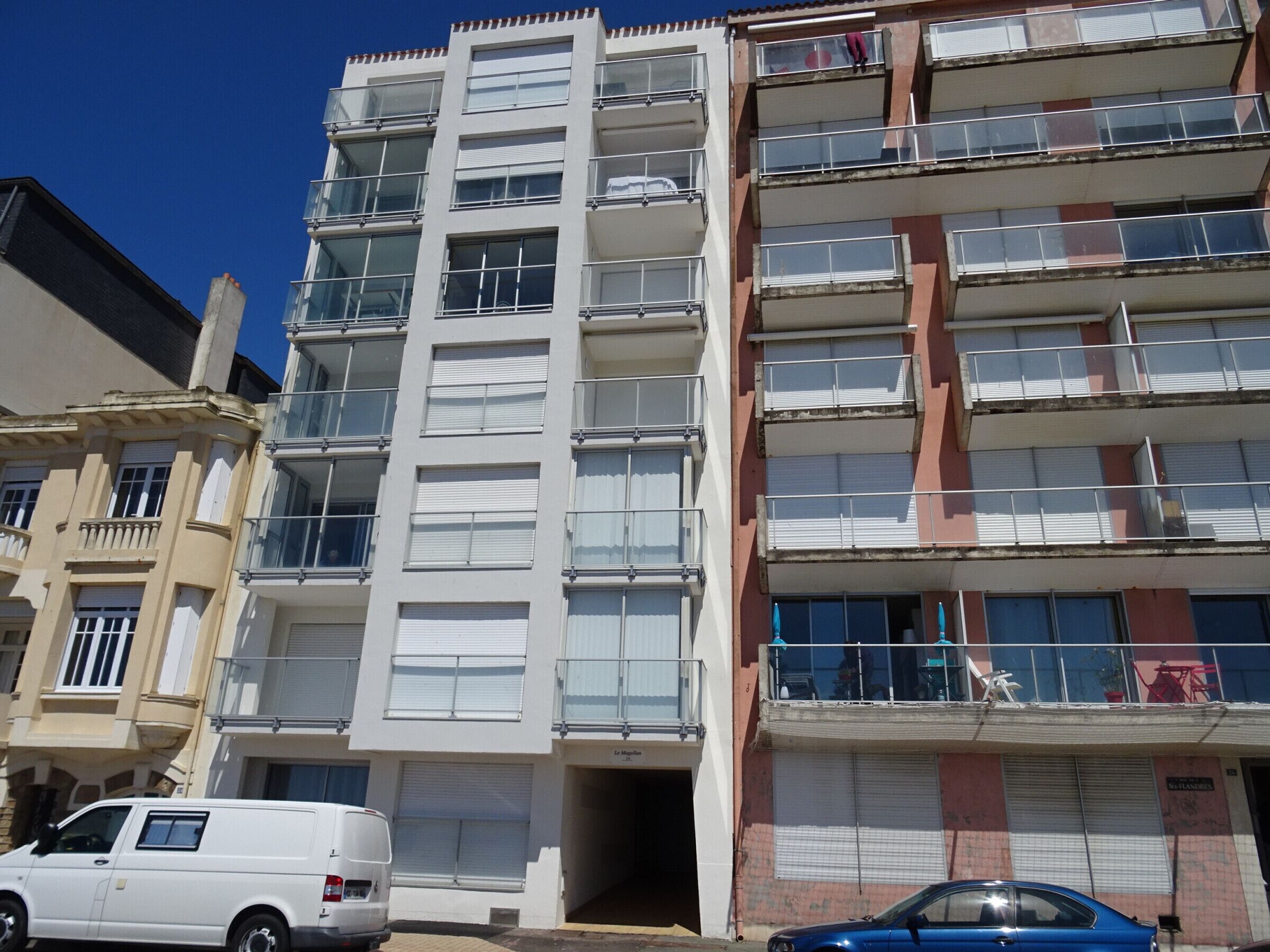 A multi-story building with balconies, a white van parked in front, and a blue car parked on the street.