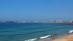 A beach with clear blue water, a sandy shore, and a city skyline in the background.