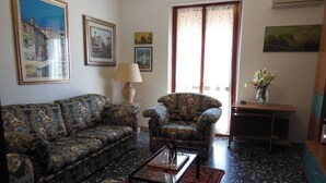 Ein Wohnzimmer mit Sofa, Sesseln und Couchtisch. An den Wänden befinden sich Bilder in Rahmen, eine Lampe und ein Fenster mit Vorhängen.