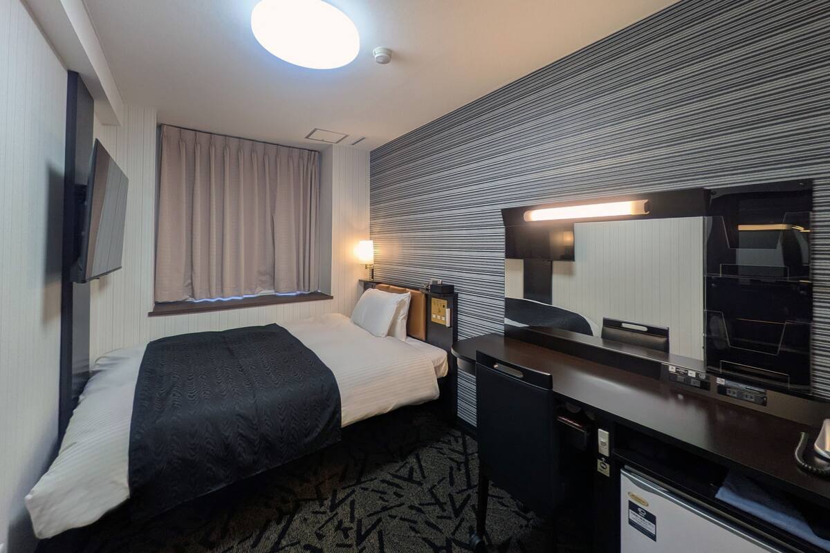 Habitación de hotel con cama, escritorio, silla, lámpara de escritorio, televisión y ventana con cortinas.