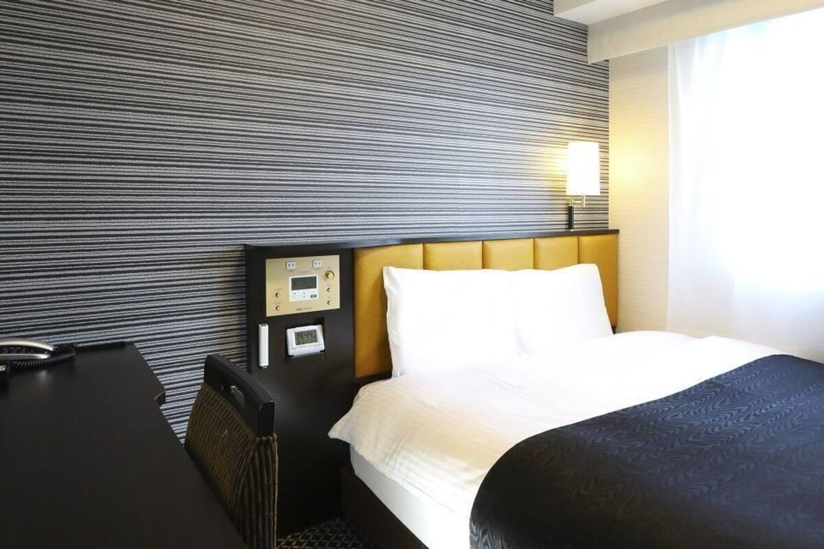 Una habitación de hotel con cama, escritorio, panel de control y ventana con cortinas.