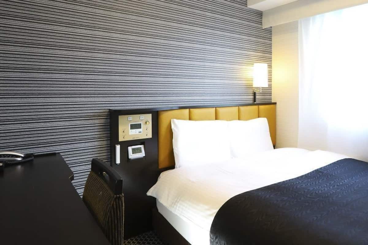 Una habitación de hotel con cama, escritorio, panel de control y ventana con cortinas.