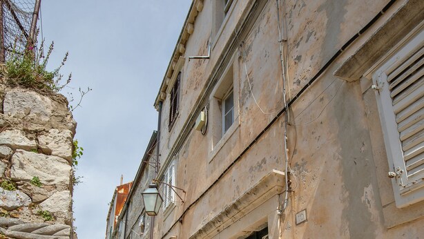 A narrow stone street flanked by historic buildings with traditional architecture, including a stone wall on one side and a building with a metal gate and decorative door. - Apartment Bell (Dubrovnik)