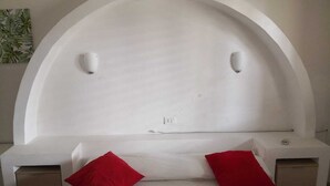 A bedroom with a white arched headboard, two bedside tables, a bed with white bedding and two red pillows, and a small nightstand with a drawer. - Residence Terrazza Vendicari (noto)