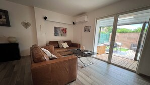 A modern living room with a brown leather sofa, a glass coffee table, and a sliding glass door leading to an outdoor patio area. - Petite Maison (Le Lavandou)