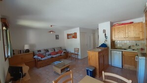 Un ambiente compatto con divano, un piccolo tavolo da pranzo, angolo cottura con pensili in legno e un frigorifero compatto.