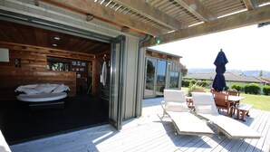 A wooden deck with a lounge area, a glass door leading to a room with a bed, and a view of the landscape. - Fantail Brand New Absolute Lakefront Whole House (Rotorua)