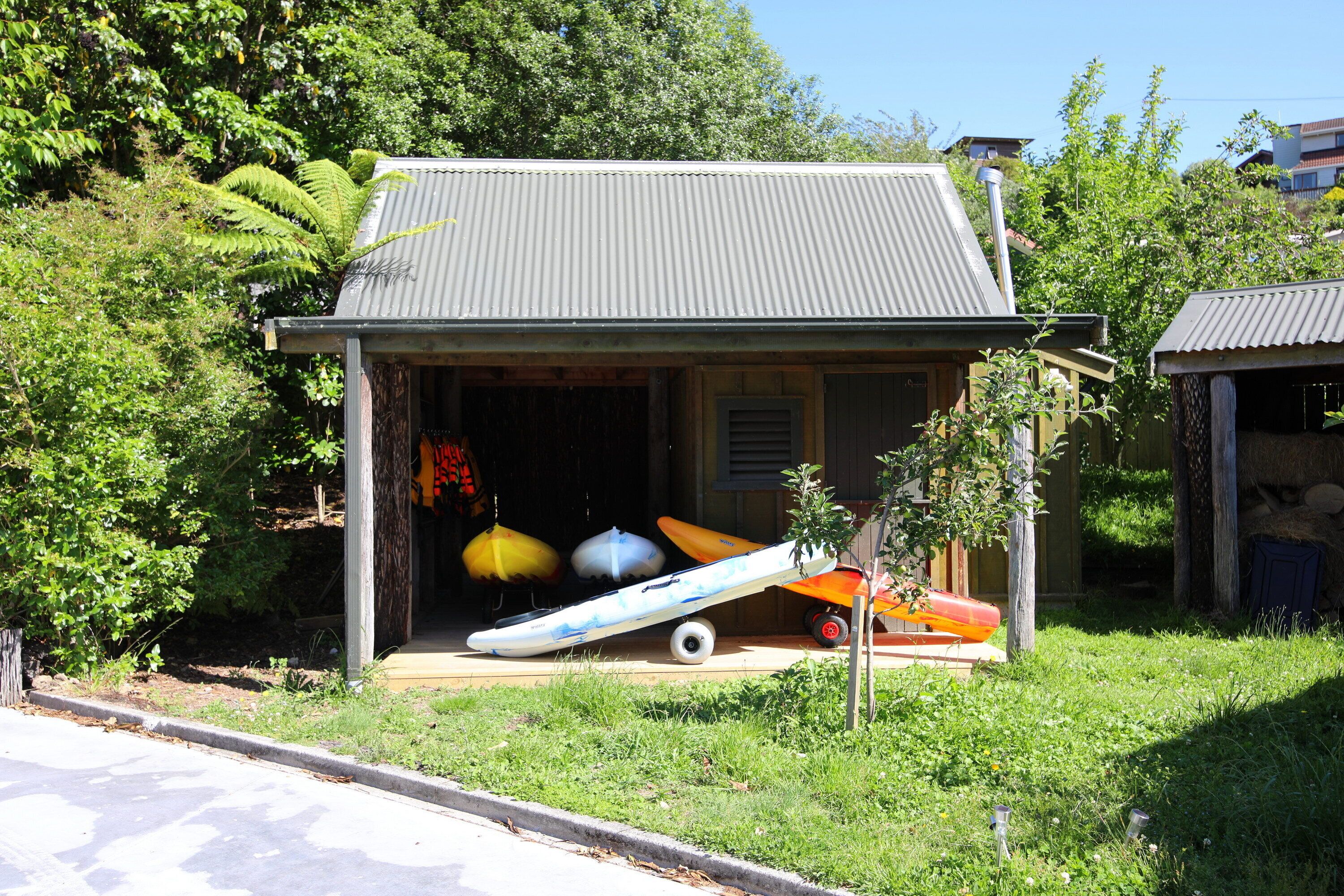 A small building with a metal roof and a door leading to a covered area. Inside, there are several kayaks and a surfboard leaning against the wall.