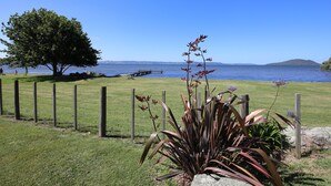 A grassy area with a wooden fence, a large tree, and a body of water in the background. - Fantail Brand New Absolute Lakefront Whole House (Rotorua)
