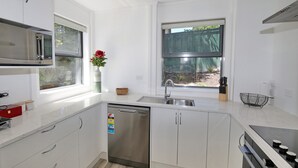 A modern kitchen with white cabinets, a stainless steel dishwasher, a sink, and a window with a view of greenery. - Fantail Brand New Absolute Lakefront Whole House (Rotorua)